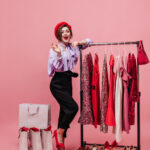 Full-length shot of elegant lady in red hat and black-and-purple outfit posing with hanger with elegant dresses, shoes and packages